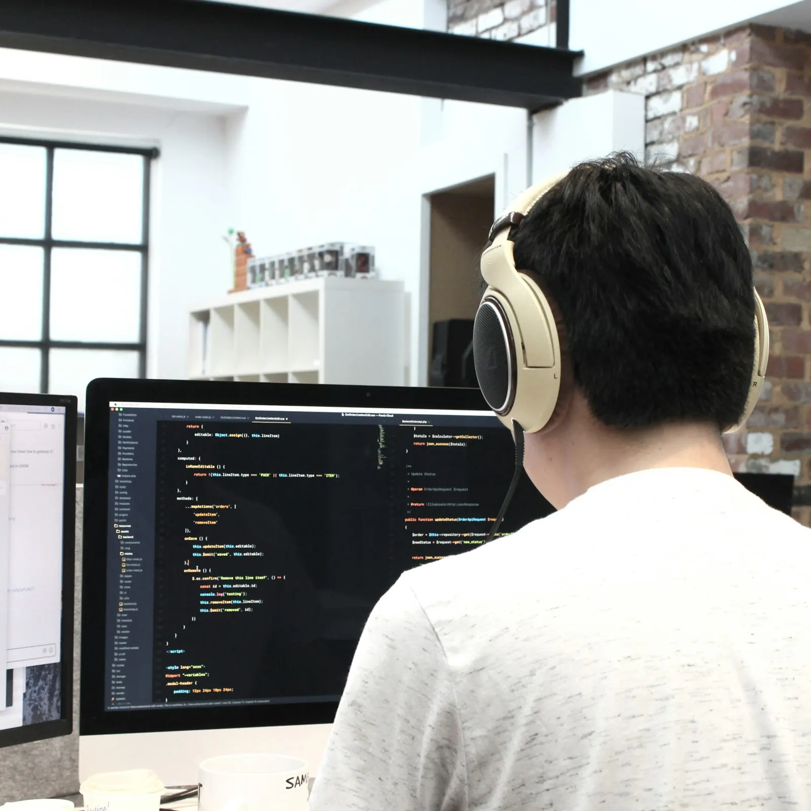 Developer memakai headphone menulis kode di depan monitor, terlihat dari belakang